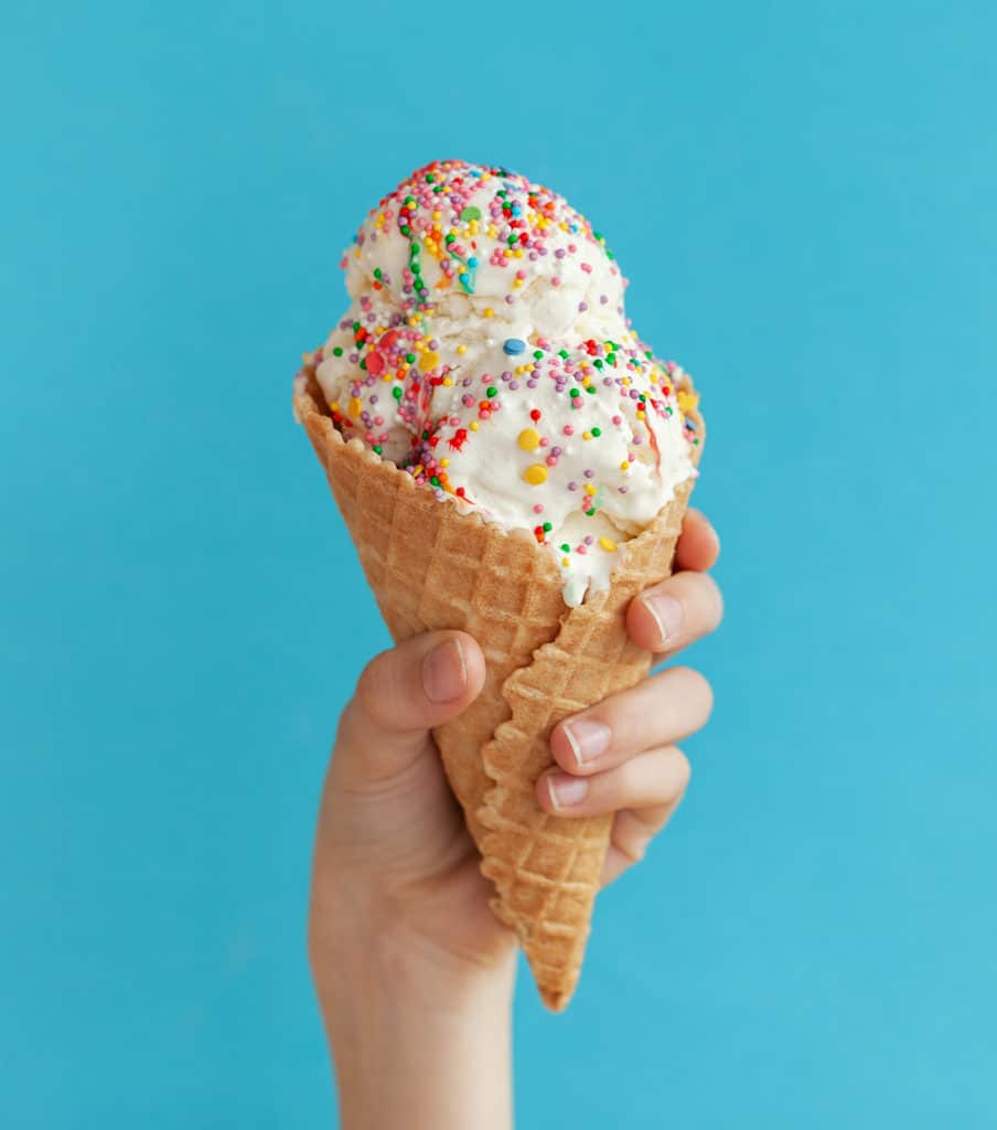 hand holding a waffle cone with vanilla ice cream covered in rainbow sprinkles for national ice cream month dairy sanitation tips